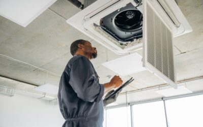 Technician in a gray uniform inspecting an office air conditioning unit and recording findings on a clipboard
